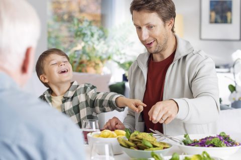 Perhe ruokapöydässä, ruutupaitainen poika ja huppariin pukeutunut isä hymyilevät ja osoittavat pinaattilettuastiaa.